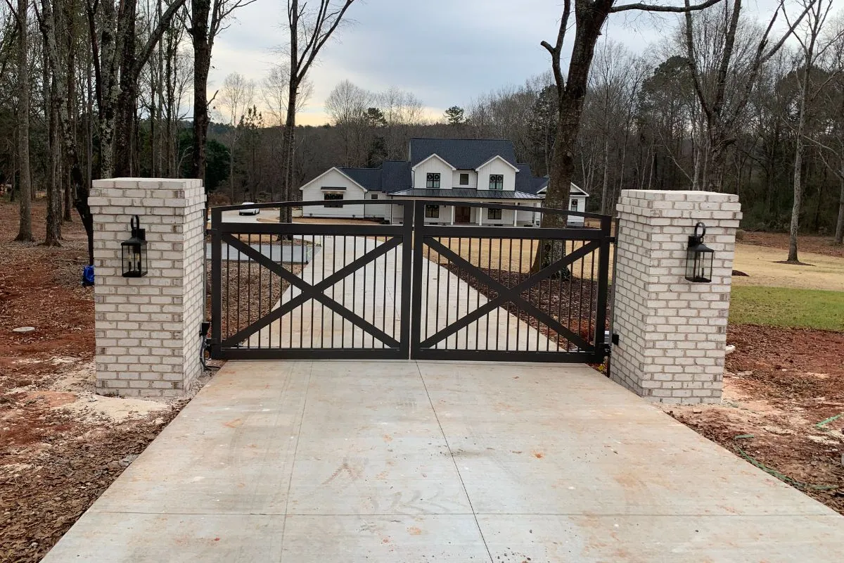 Custom arched aluminum driveway gate with X-brace panels, set between white brick columns at a modern farmhouse entry